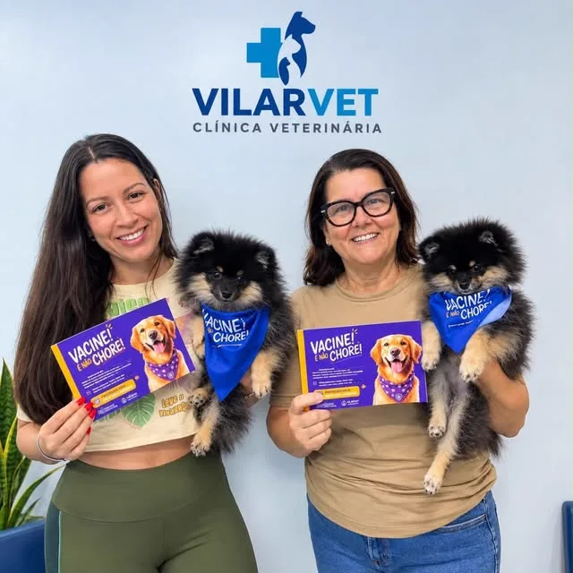 Duas profissionais da VilarVet seguram dois filhotes de spitz com bandanas azuis e a cartilha de vacinação.