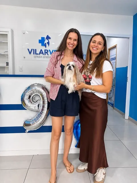Duas profissionais na recepção da VilarVet com um shih-tzu no colo e um balão de número nove, no aniversário da clínica.